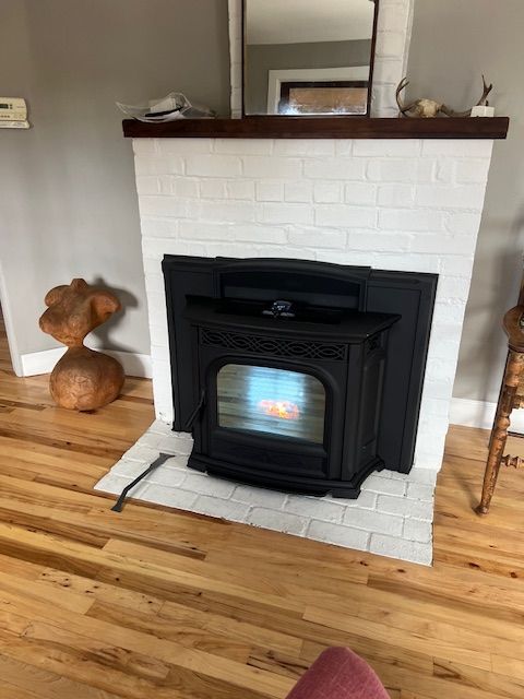 A black pellet stove sits inside a white-painted brick fireplace, with a fire burning inside. The floor is hardwood.