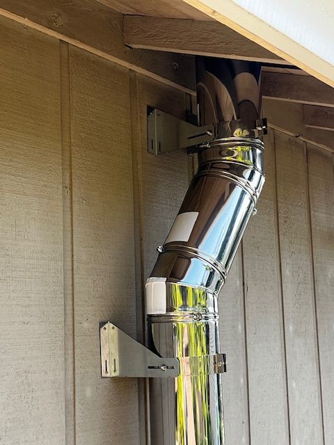 A shiny metal chimney pipe is mounted on the side of a wooden building, secured by metal brackets. The pipe bends at an angle.