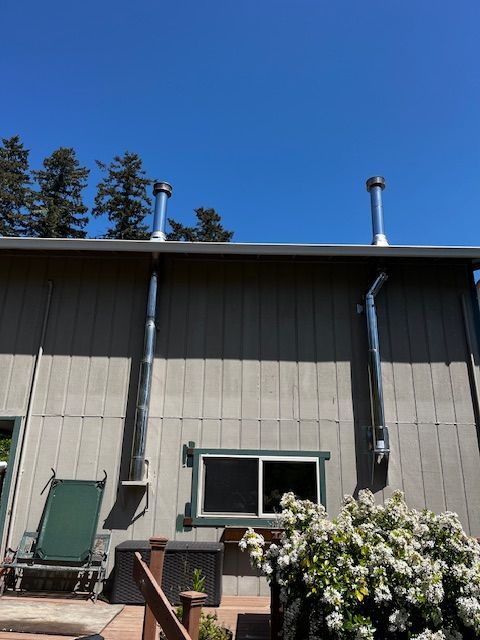 Two shiny metal chimneys extend from a building's roof, against a blue sky. The building is light grey with a small window and a deck.