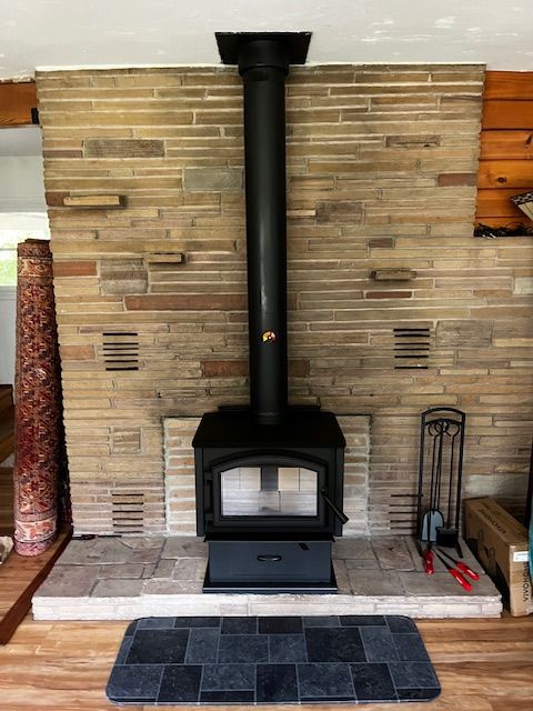 A black wood-burning stove with a tall pipe against a tan stone wall. A mat and tools sit in front of the stove on a tile hearth.