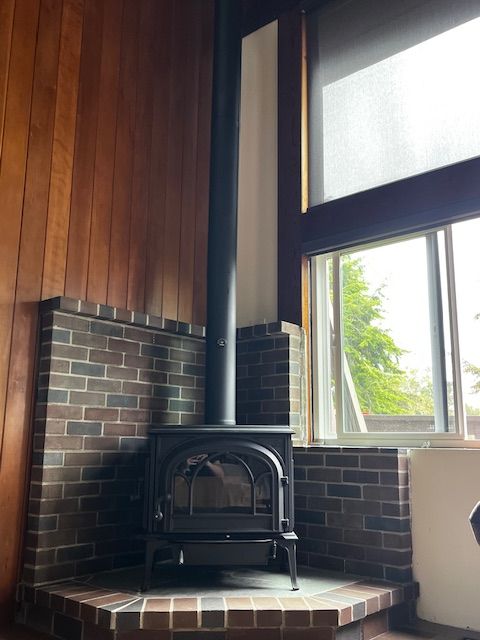 Wood-burning stove with black chimney against brick and wood paneling, next to a window.