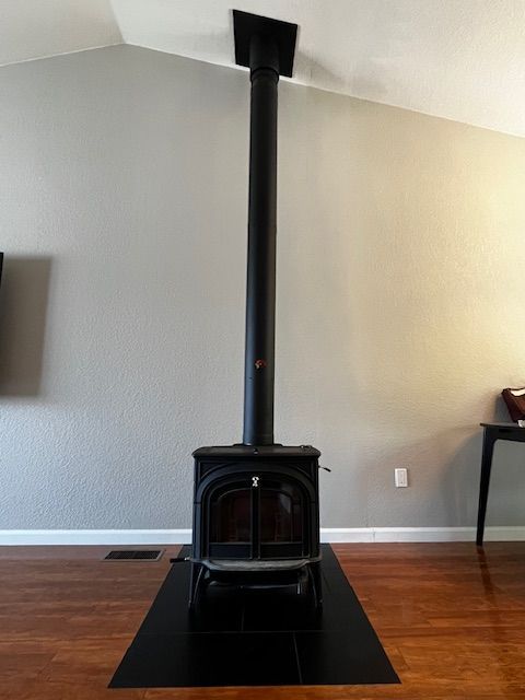 A black wood-burning stove with a tall flue pipe against a light gray wall. It sits on a black tile hearth in a room with wood flooring.