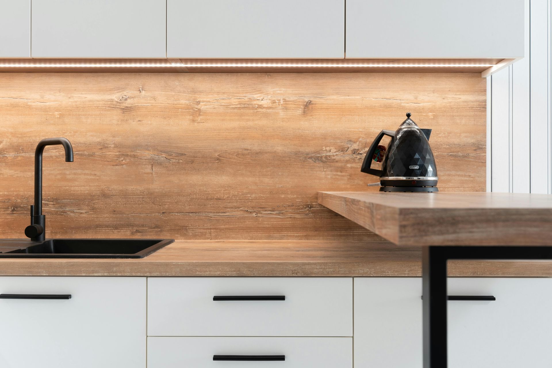 Modern kitchen with black sink, faucet, and kettle against wood grain backsplash and white cabinets.