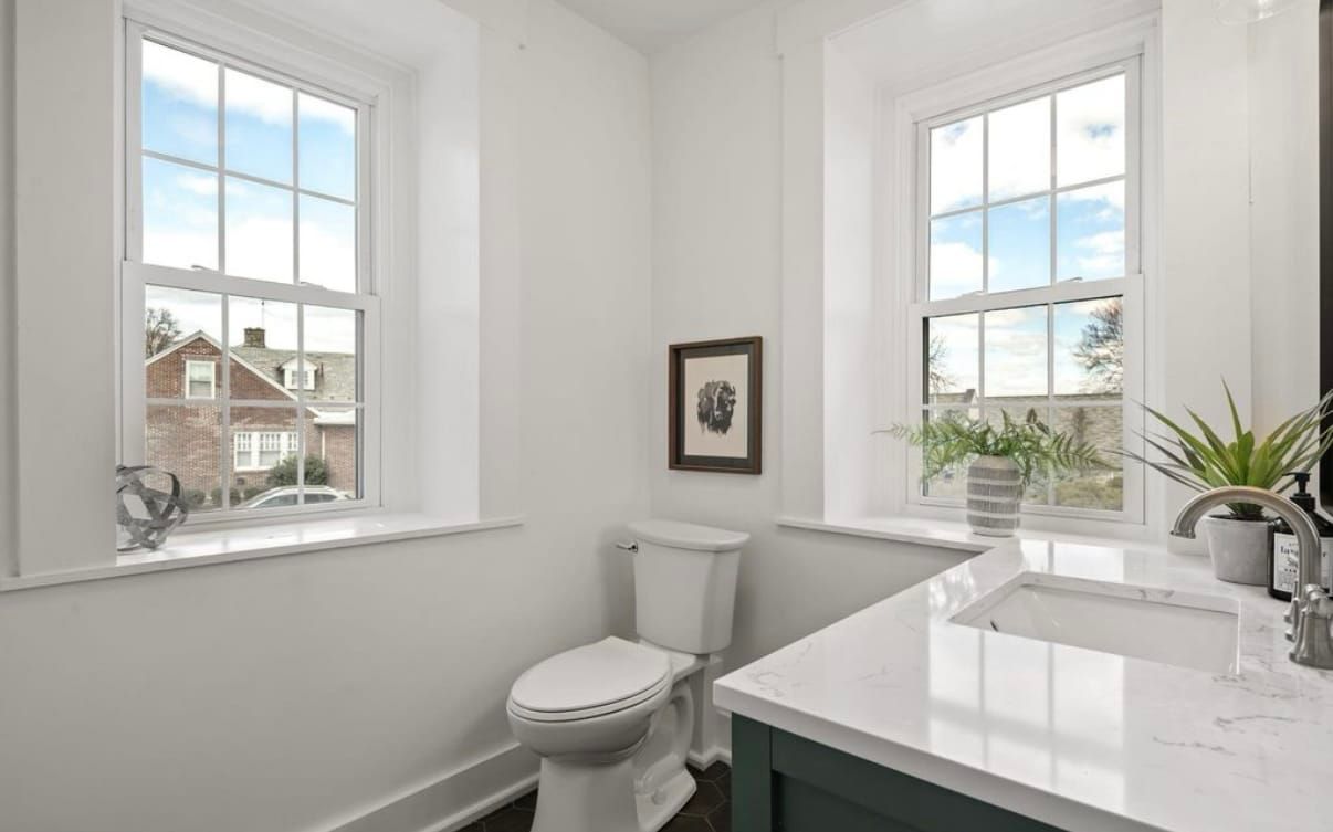 A bathroom with a toilet , sink and two windows.