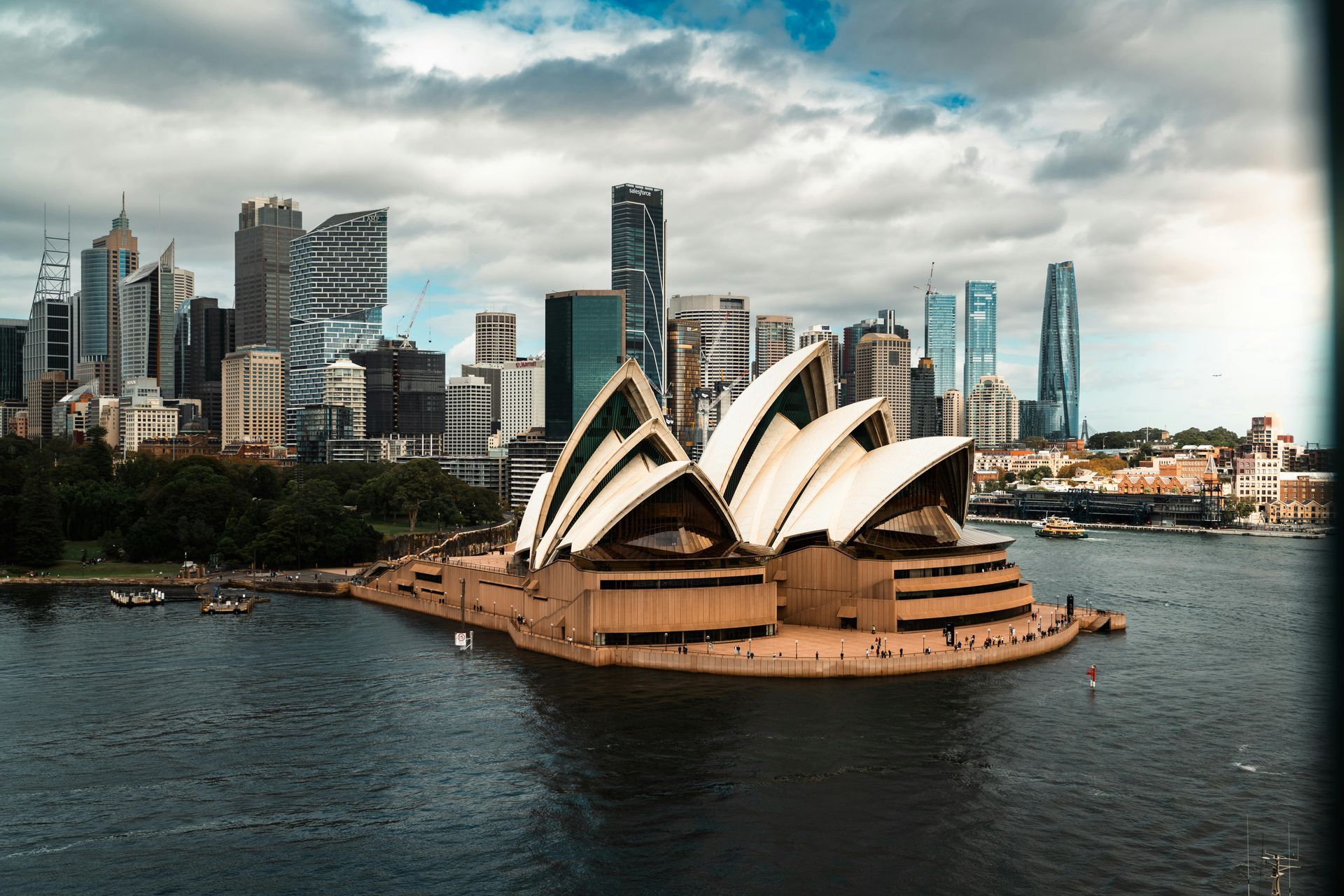 Sydney Opera House on the harbor with city skyscrapers under a cloudy sky