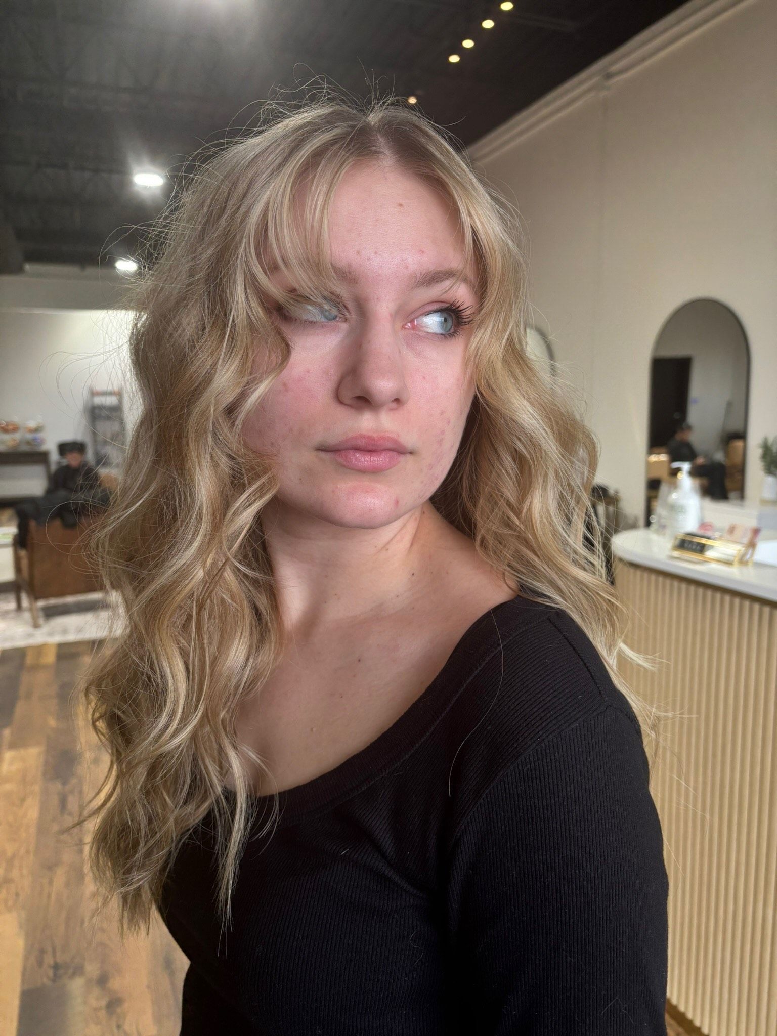 Blonde-haired person with wavy hair, in a black top, looks off-camera in a salon.