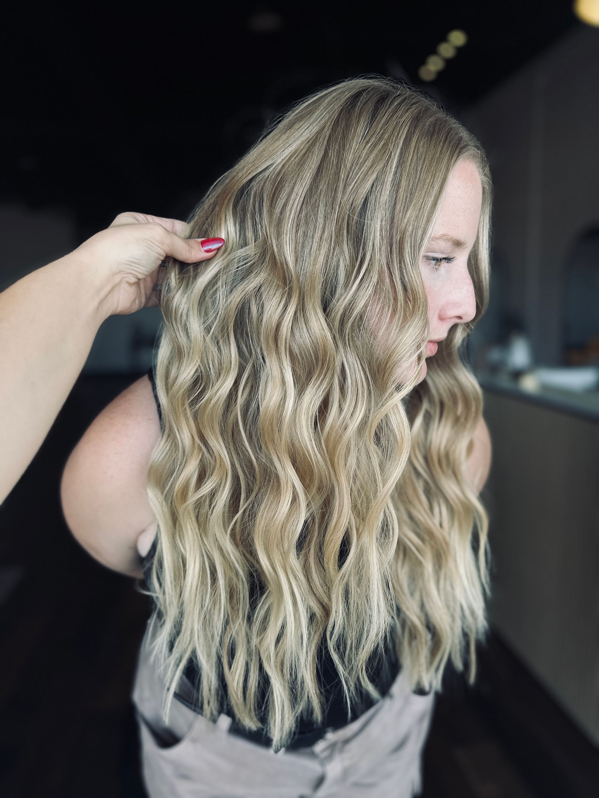 Woman with long, wavy blonde hair; hand touches hair. Indoor setting, soft lighting.