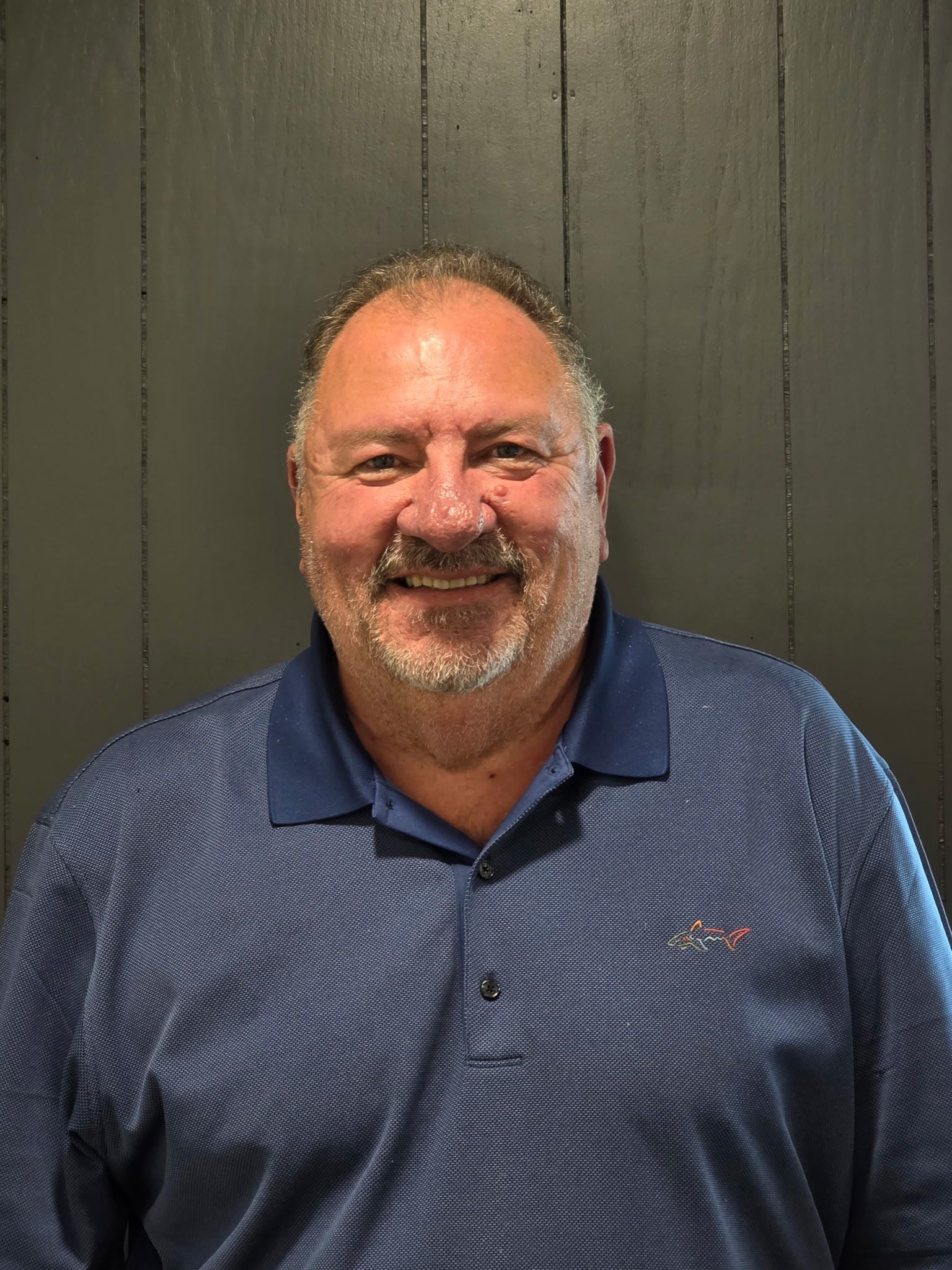 A smiling person with a beard, wearing a blue patterned polo shirt in front of a dark vertical wooden panel wall.