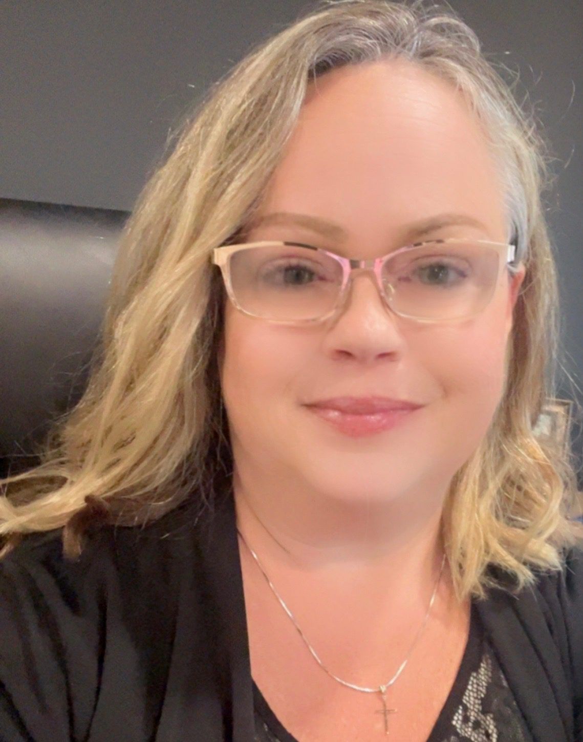 A person with shoulder-length blonde hair wearing glasses and a cross necklace, smiling in front of a dark background.