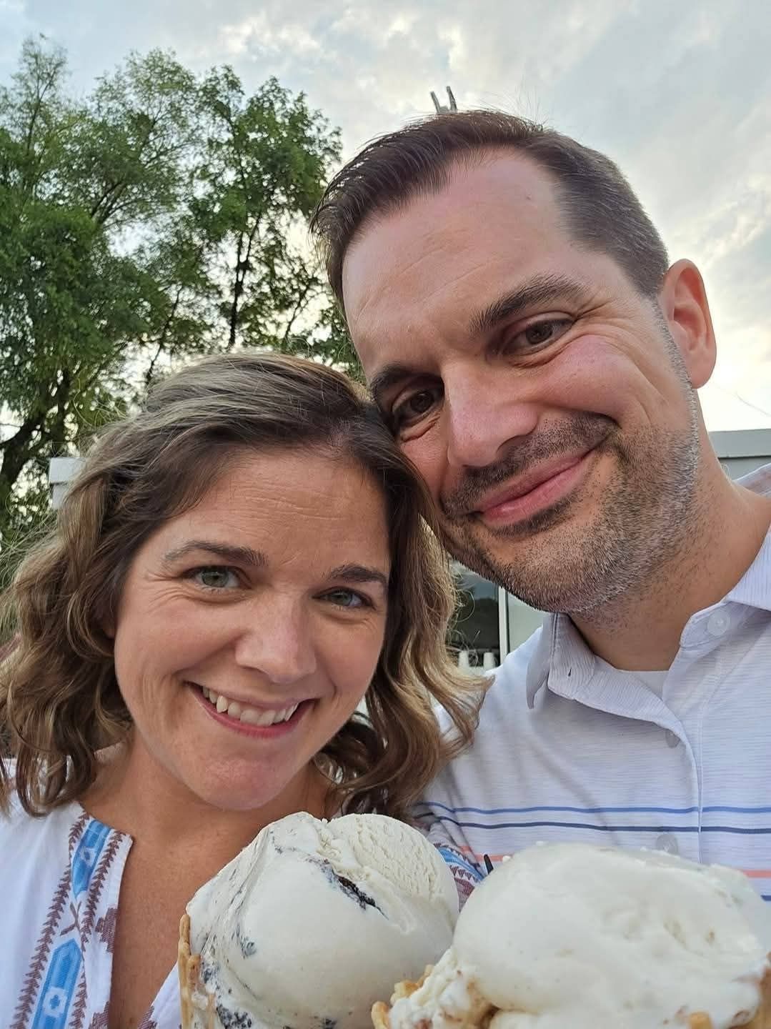 A couple smiling outdoors while holding large scoops of ice cream in waffle cones.