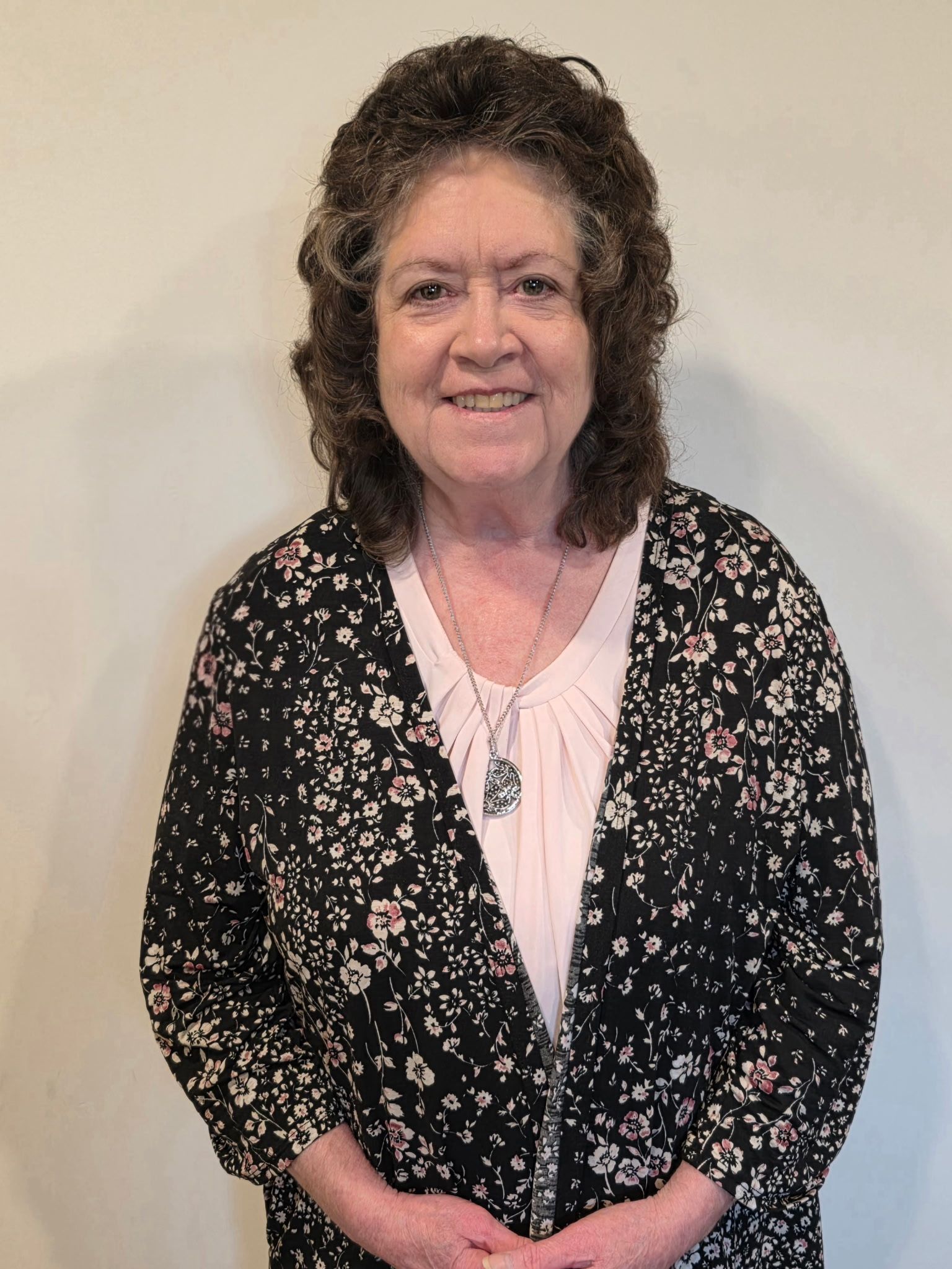 Smiling person with shoulder-length curly brown hair wearing a dark maroon cowl-neck top standing near a wooden door.