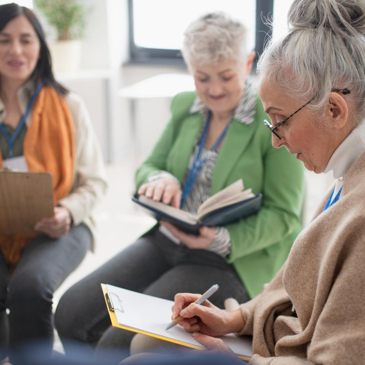 Three people sitting in a circle, one writing on a clipboard, another reading a book, and the third holding a clipboard.