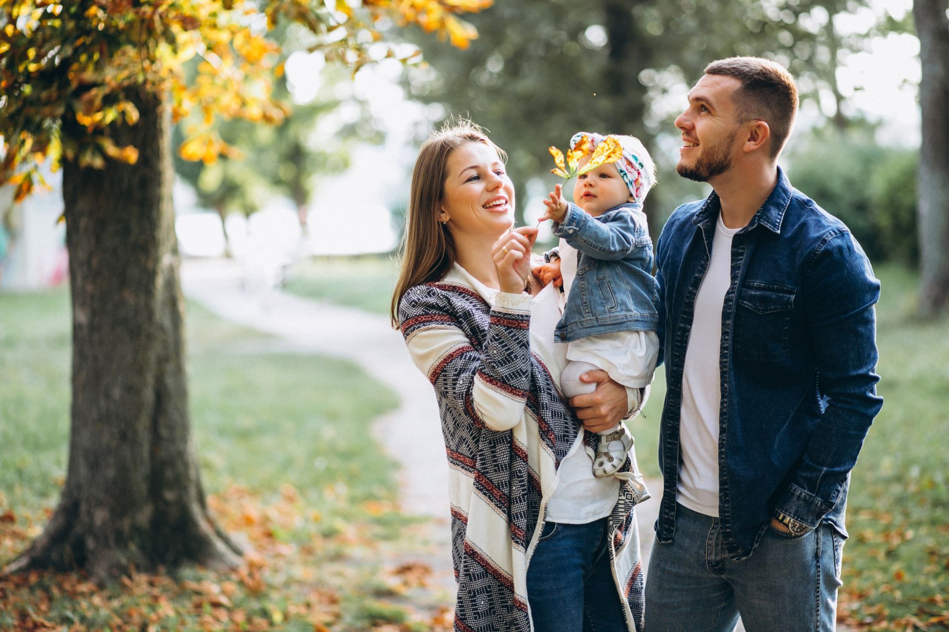 A family stands in a park during autumn; a parent holds a child who reaches toward yellow leaves while another looks on.