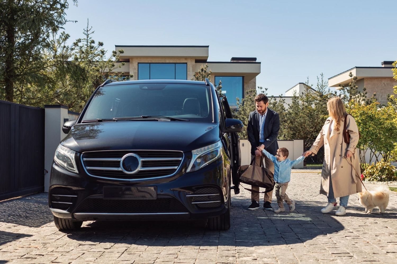 A family with a small dog prepares to board a black minivan parked in front of a modern home on a sunny day.