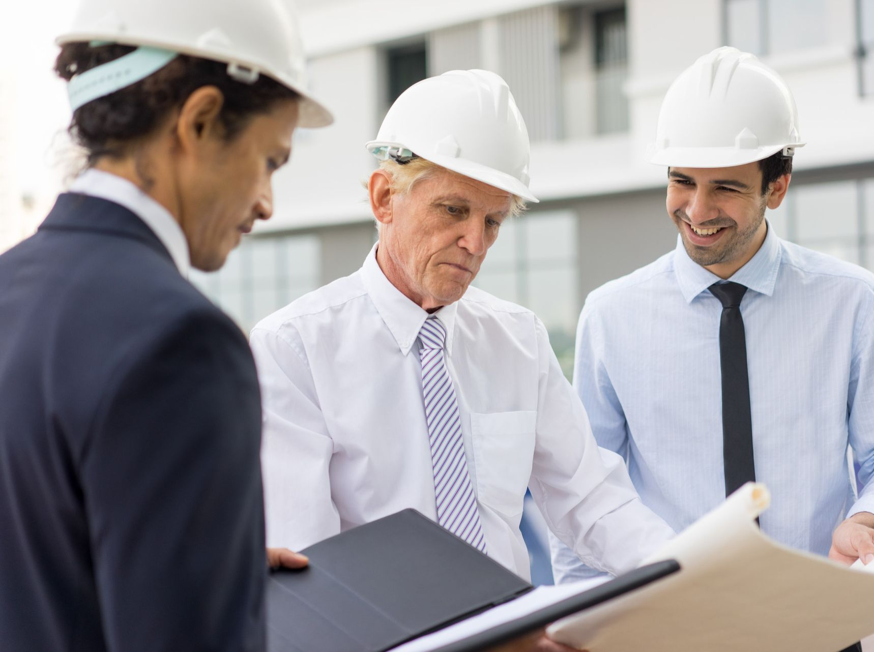 Three professionals in hard hats and business attire review construction blueprints together outdoors.