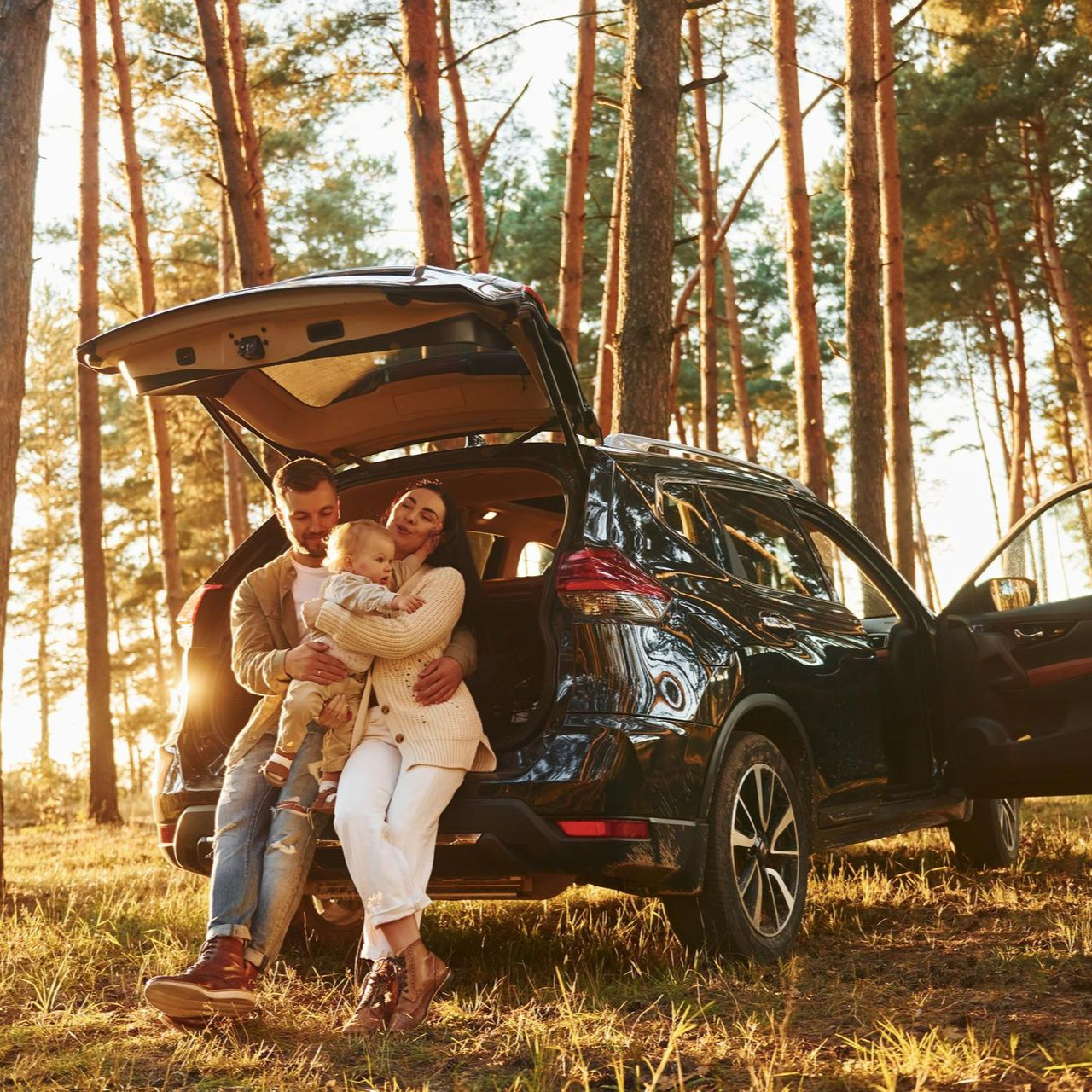 A family sits in the open trunk of a dark SUV parked in a sunlit pine forest at golden hour.