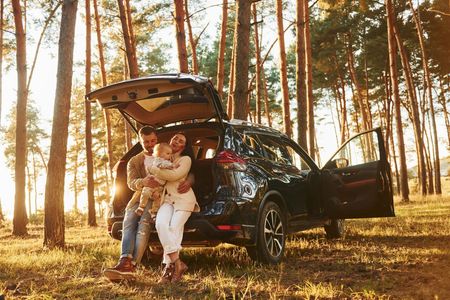 A family sits in the open trunk of a black SUV parked in a sunlit pine forest.