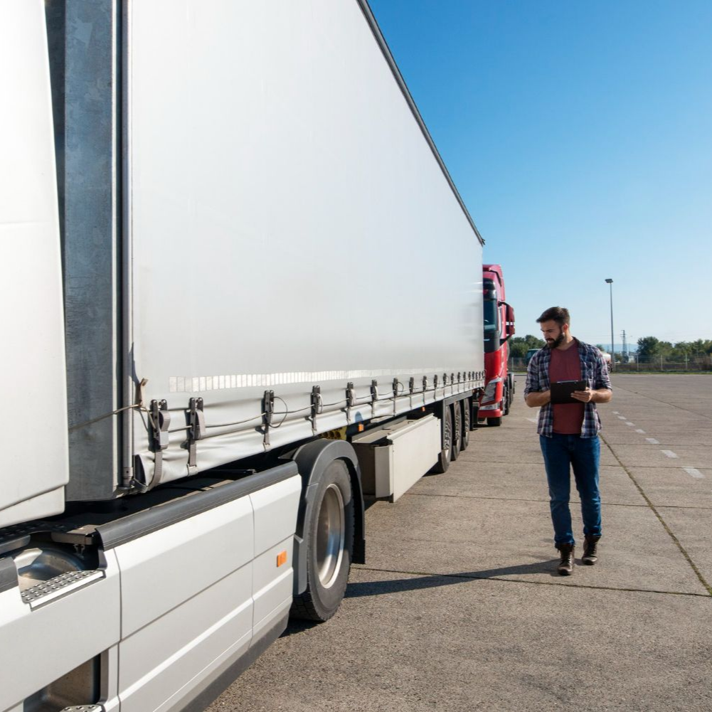 A person walks beside a large semi-truck in an outdoor parking lot while looking at a digital tablet.