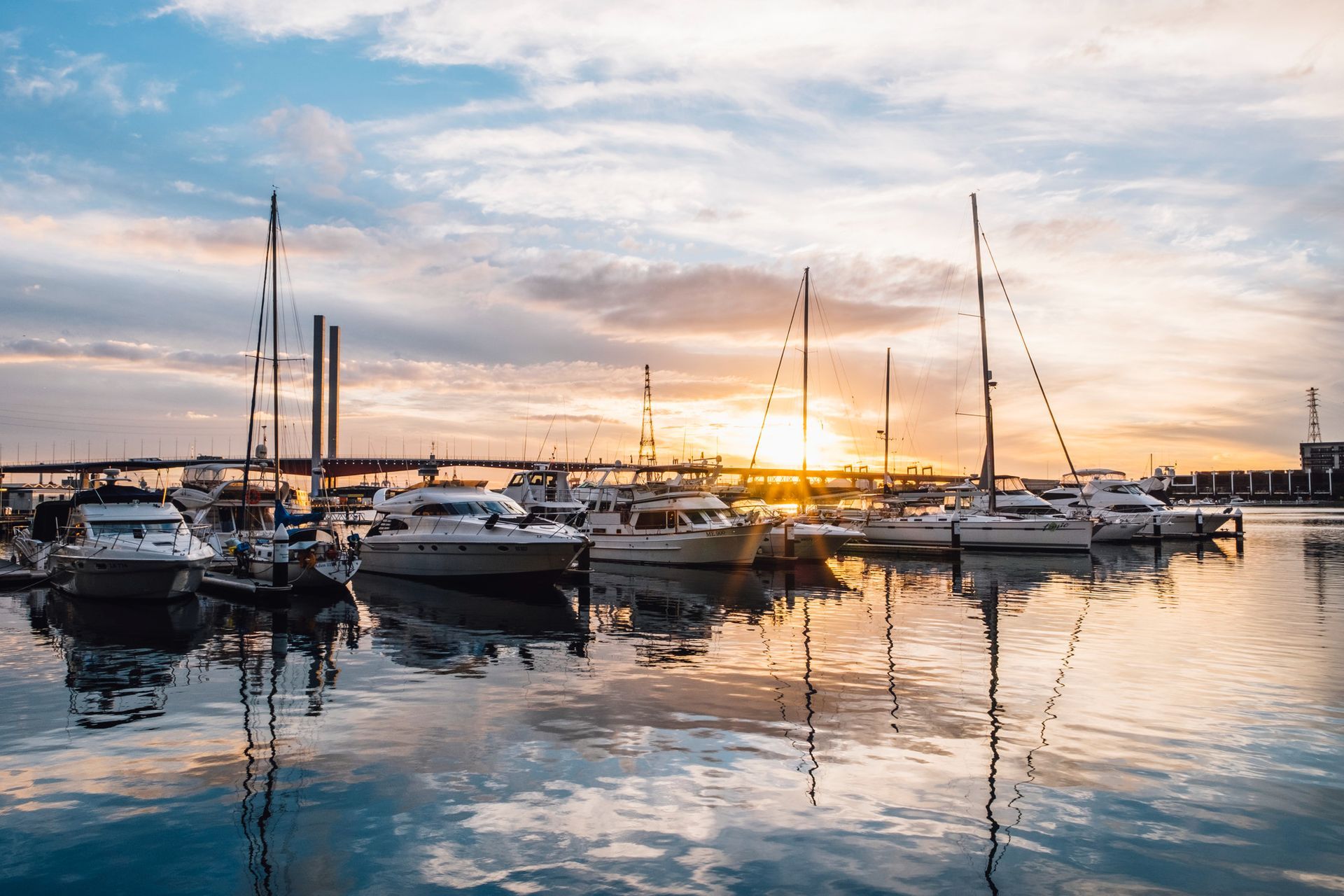 Boats docked in a harbor during a golden sunset, with reflections on the calm water and a bridge in the distance.