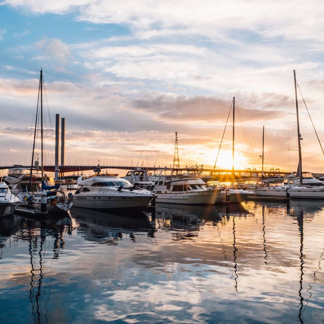 A marina filled with sailboats and yachts at sunset, with a bridge in the background reflecting on calm water.