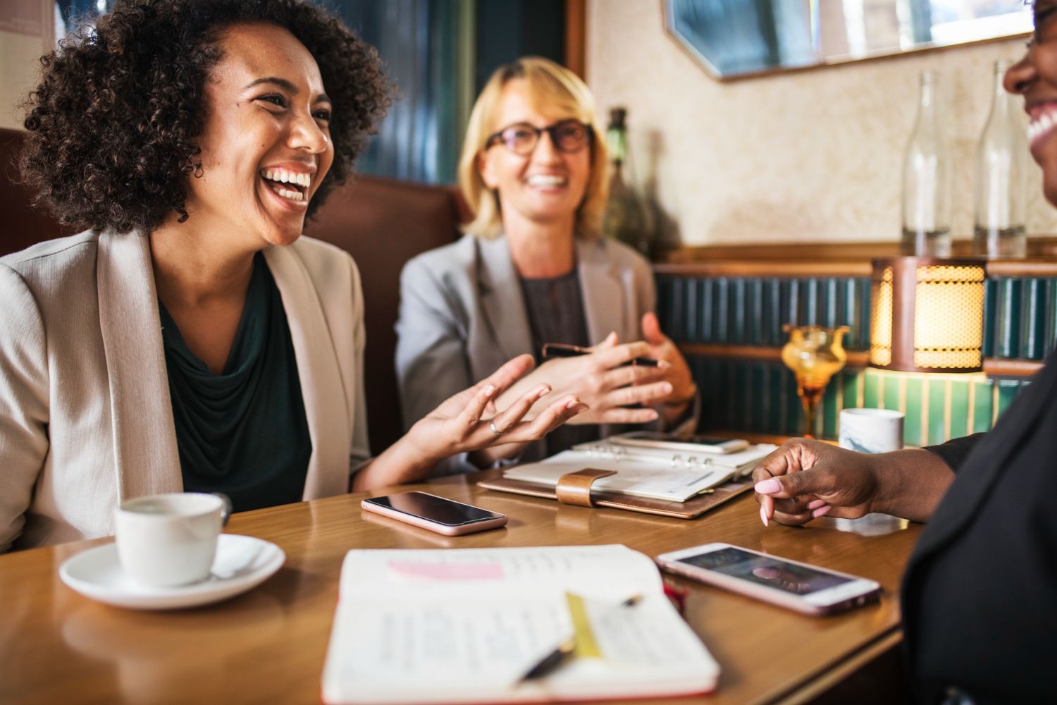 Three people in professional attire laugh during a collaborative meeting at a table with notebooks and smartphones.