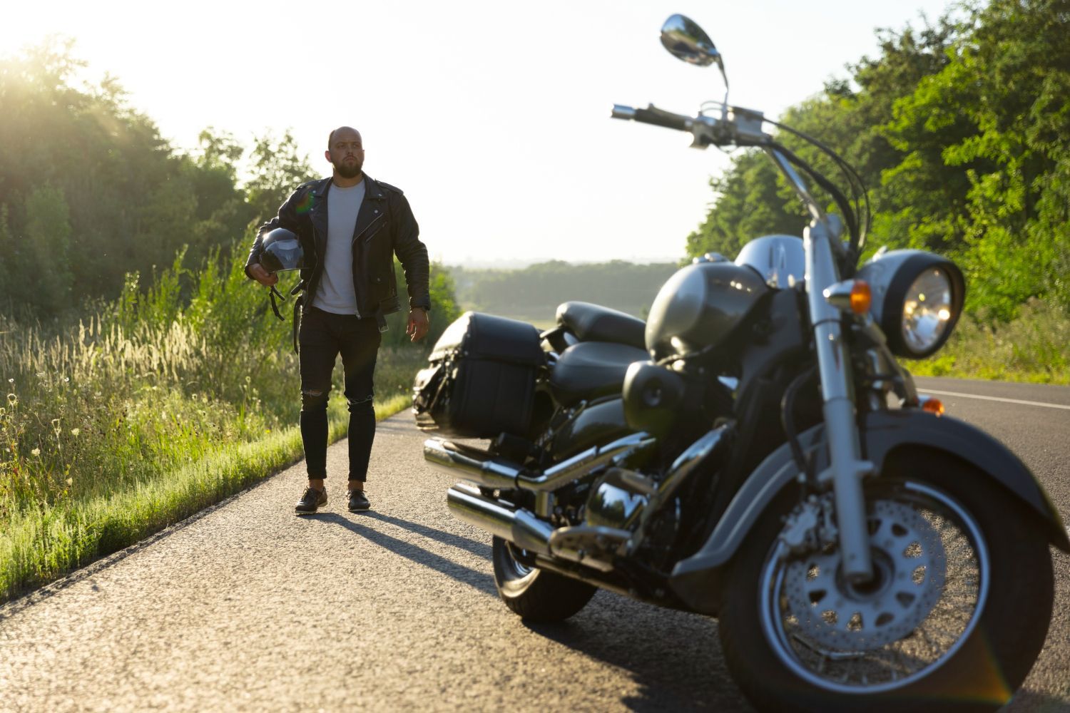 A person holding a helmet walks toward a cruiser motorcycle parked on a sunlit road bordered by trees.