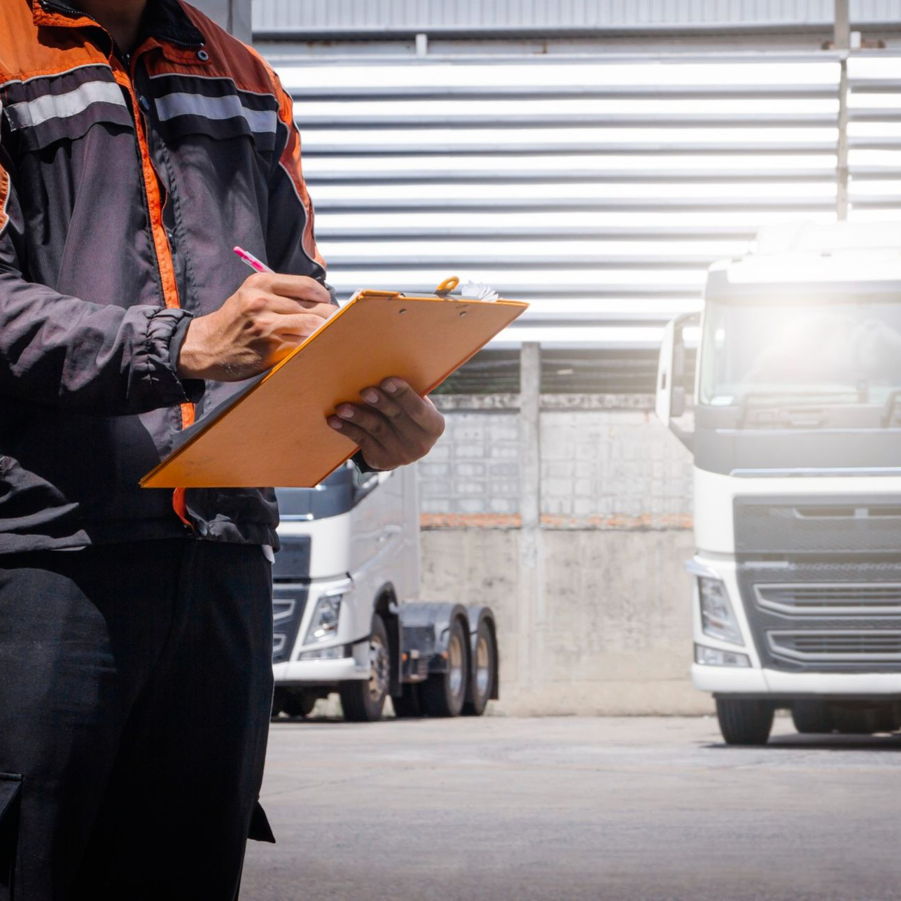 A person in a work uniform writes on a clipboard with a truck in the background at an outdoor facility.