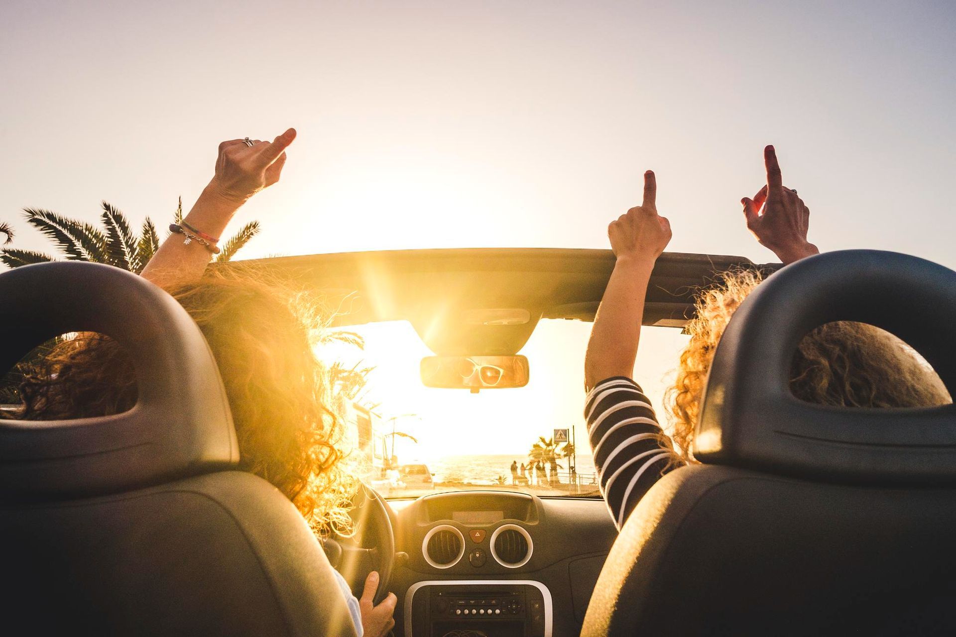 Two people in a convertible car driving toward a bright sun with their arms raised in celebration.