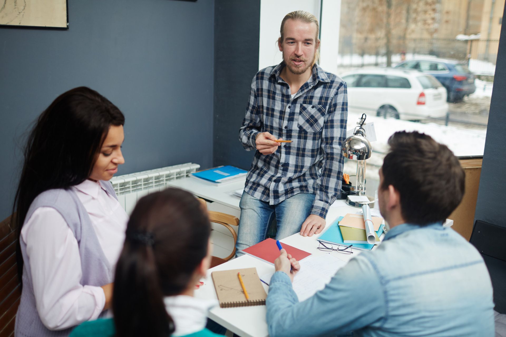 A person in a plaid shirt stands by a window, speaking to two colleagues seated at a table during a meeting.