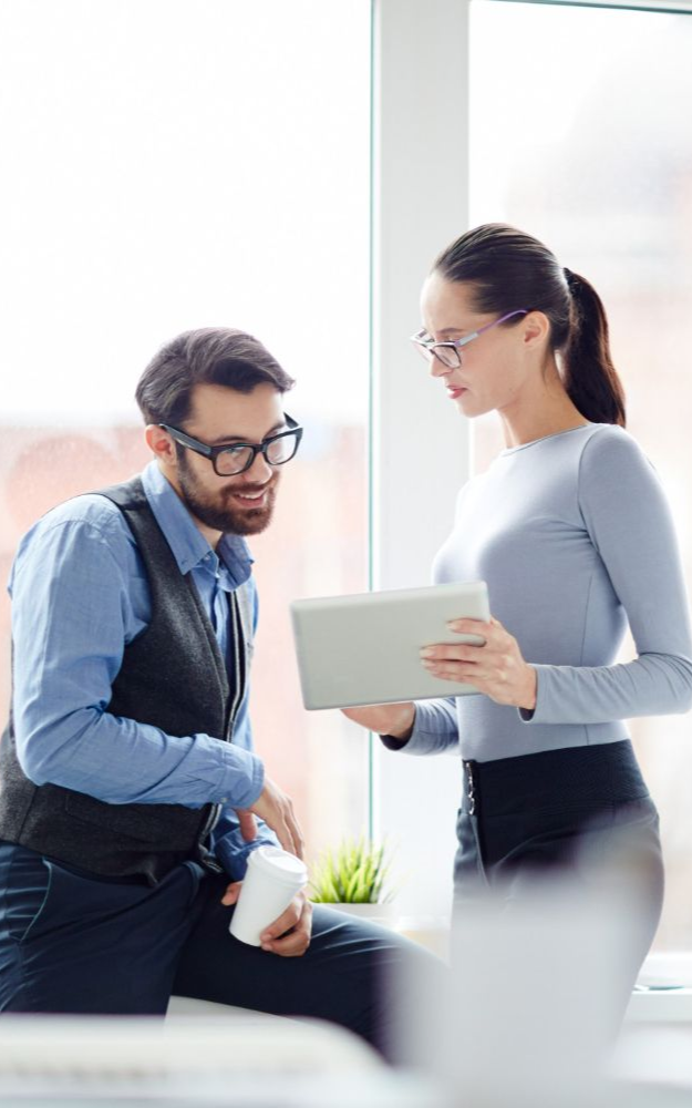 Two professionals in business casual attire stand near a window, reviewing information together on a tablet.