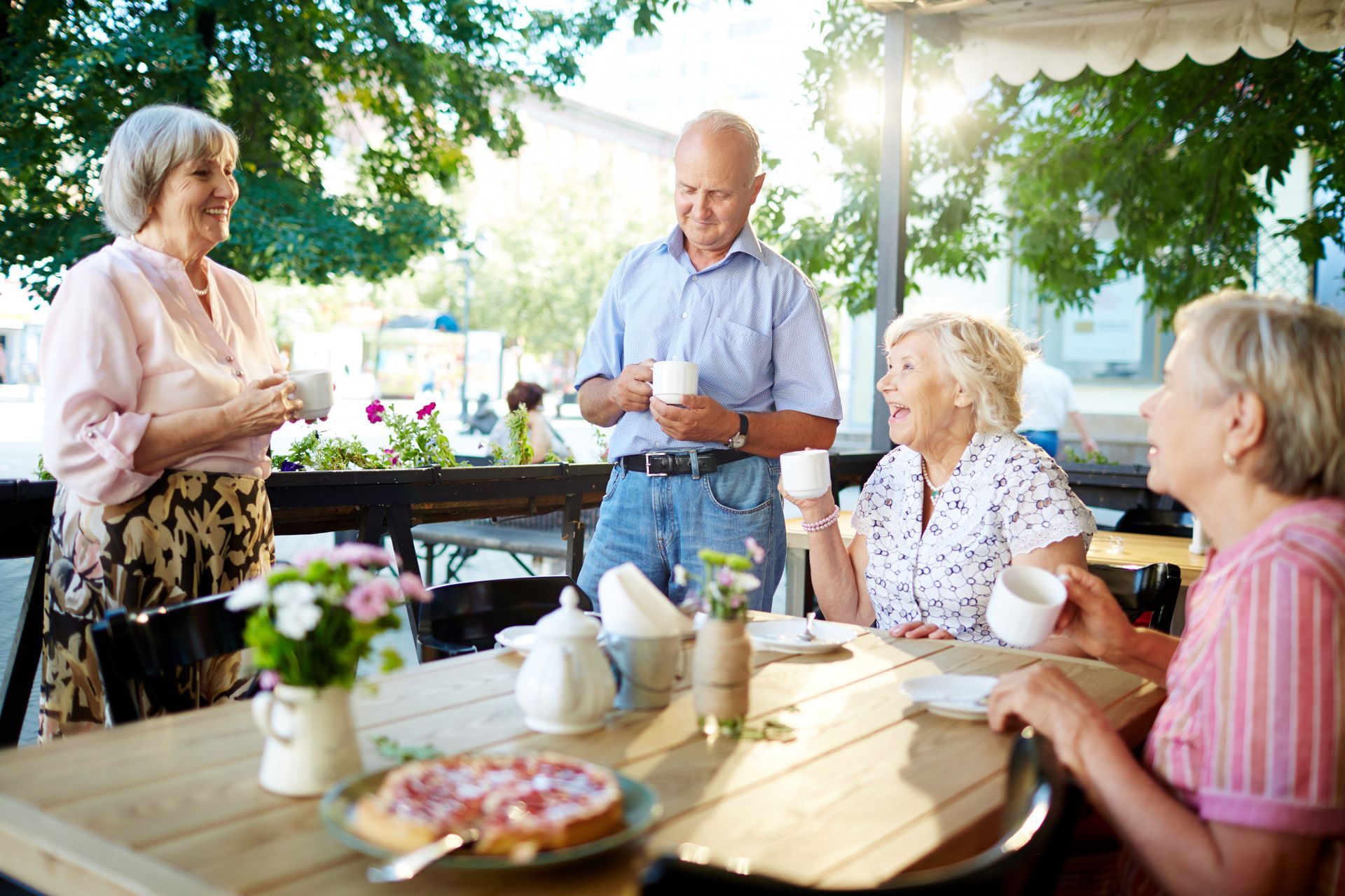 A group of people chat and drink tea at an outdoor wooden table with a pizza, set under trees in sunny, dappled light.