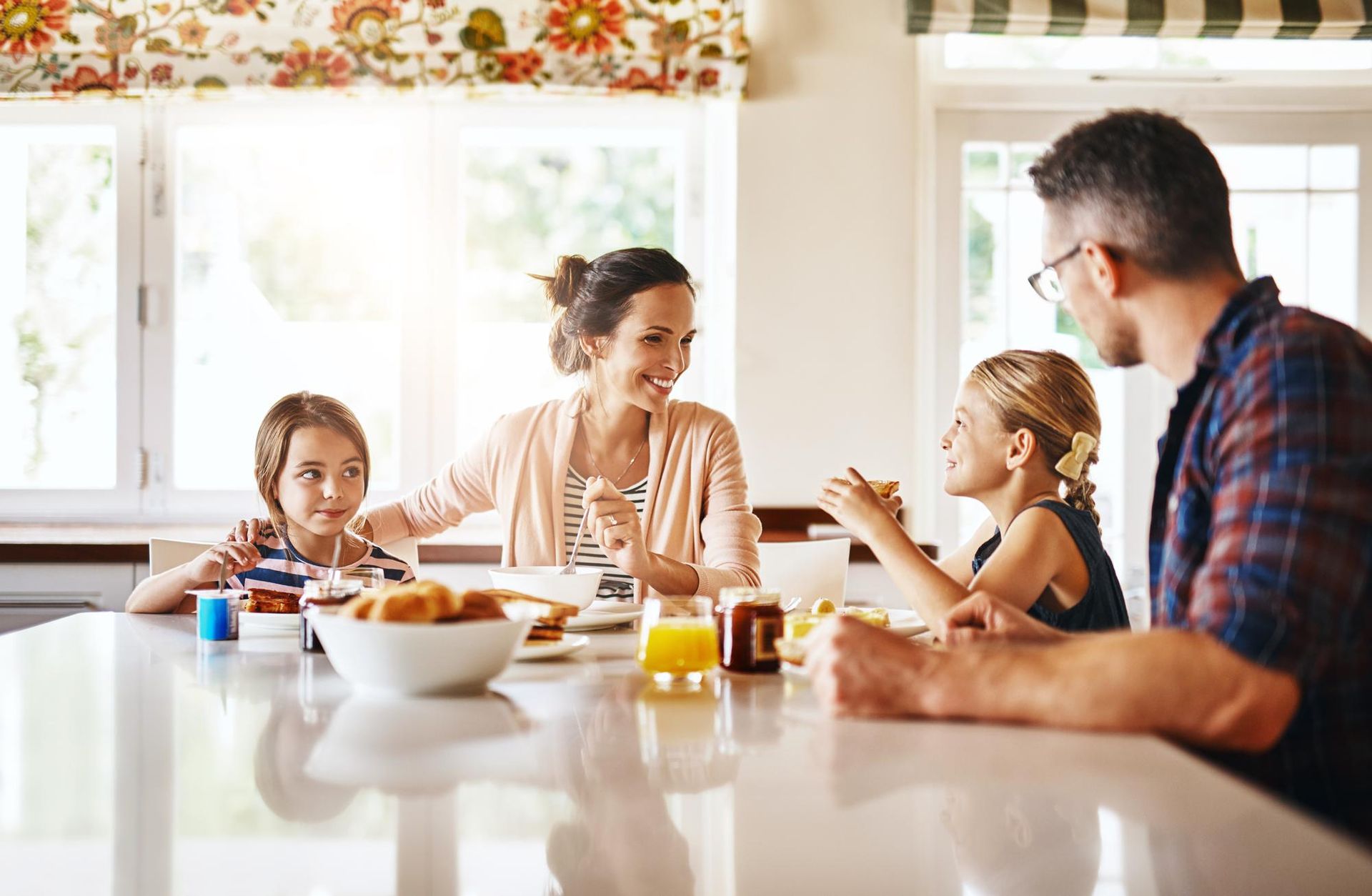 A family sits at a bright kitchen counter, smiling and talking while eating breakfast in a sunlit room.