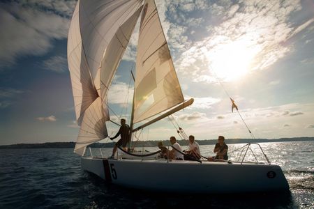 A sailboat with white sails travels across a sunny, sparkling sea with four people on deck.