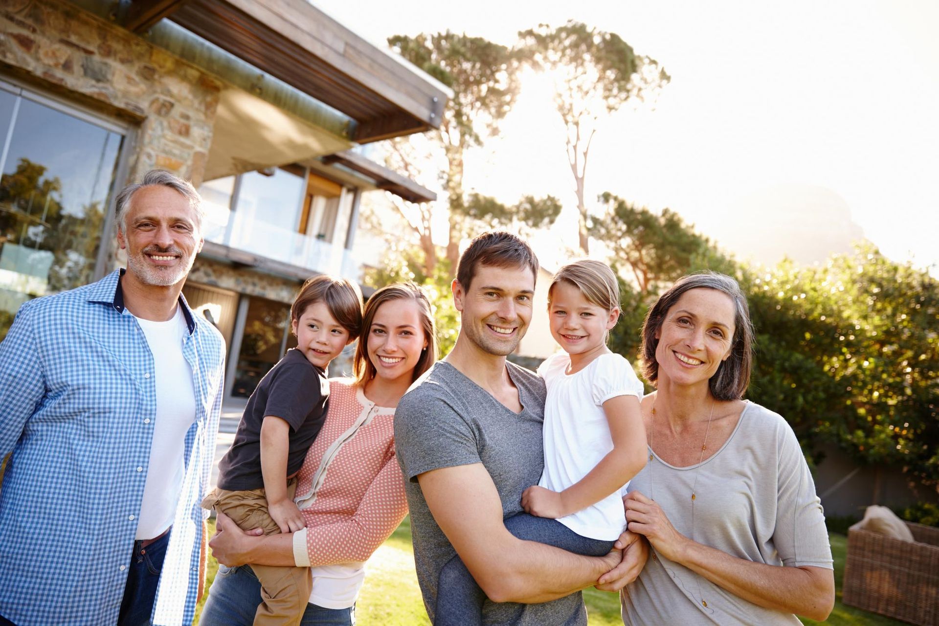 A multi-generational family smiling together outside their modern home on a sunny day.