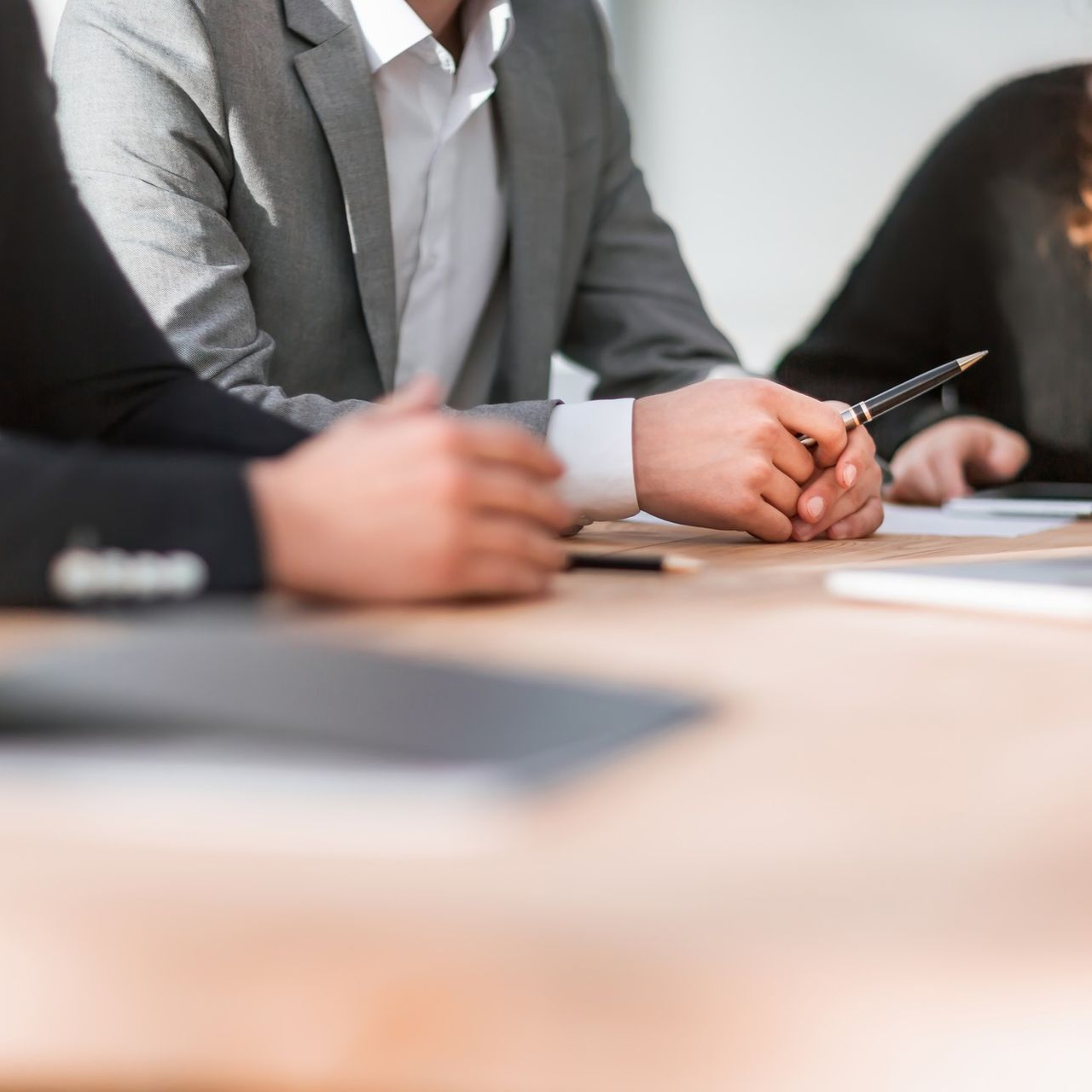 Business professionals in formal attire sit at a table in a meeting, with hands resting on the surface.