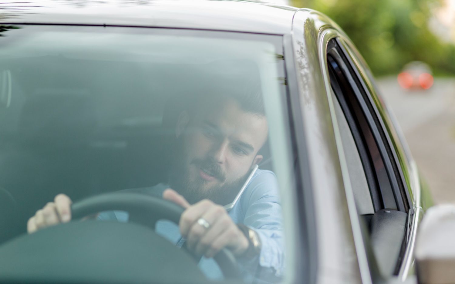 A person with a beard driving a car while talking on a mobile phone held to their ear.