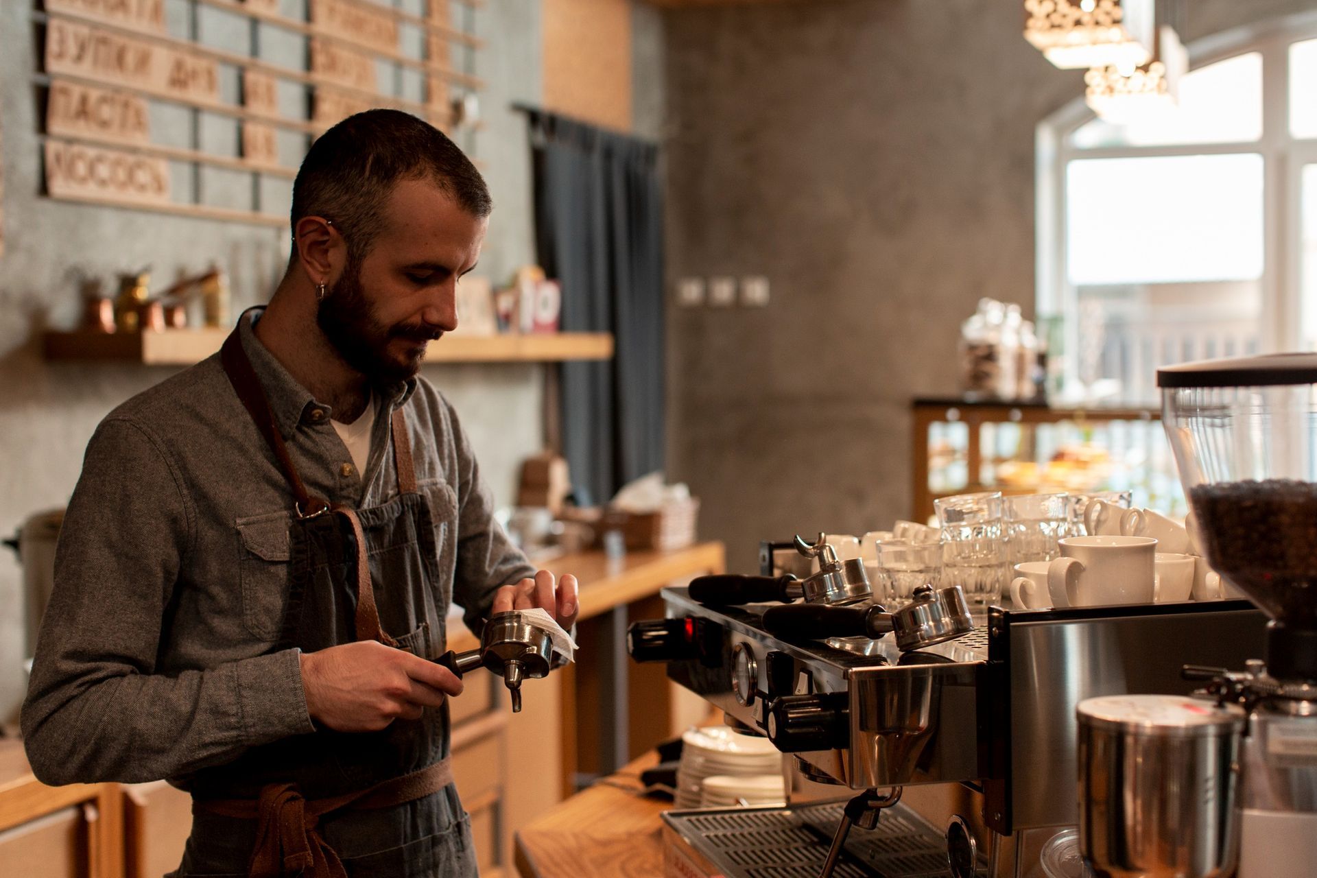 A bearded barista in a brown apron prepares a portafilter at an espresso machine in a modern, warm-toned cafe.