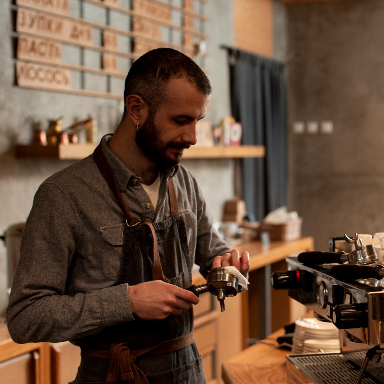 A barista in a gray shirt and apron preparing espresso with a portafilter behind a coffee shop counter.