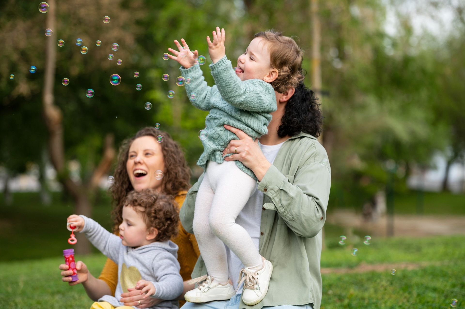 A family plays with bubbles in a park; a child is held up while reaching for bubbles as another child holds a bubble wand.