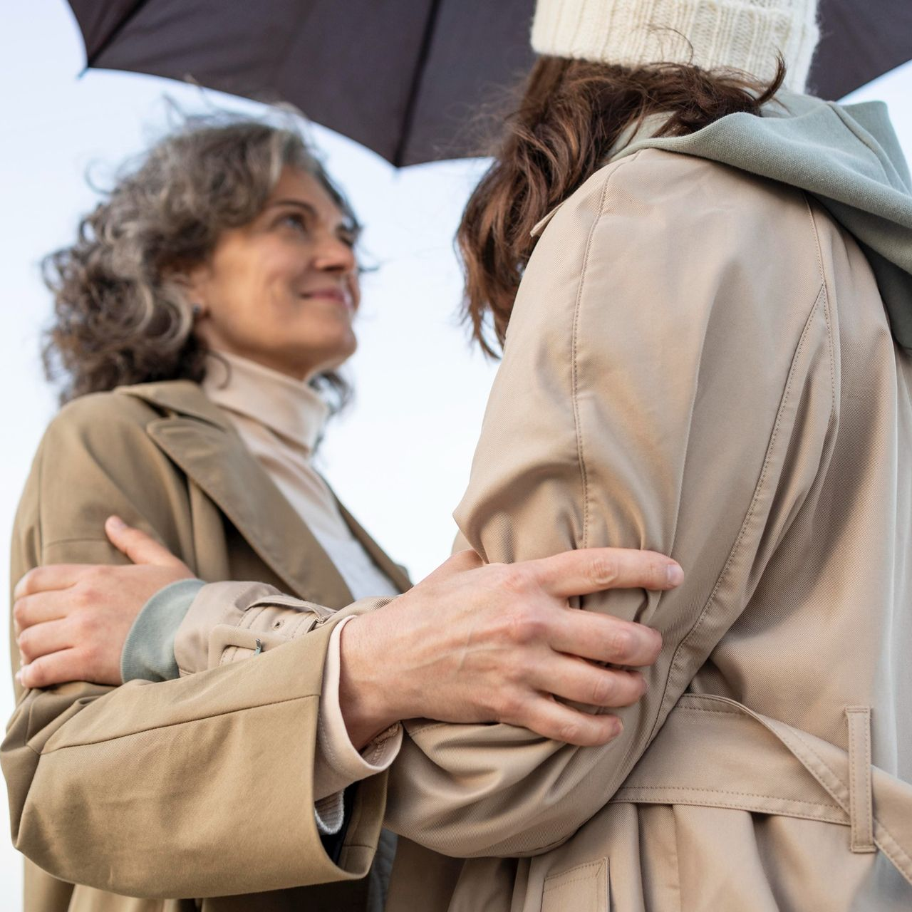 Two people in beige trench coats embrace under an umbrella, with one looking fondly at the other.