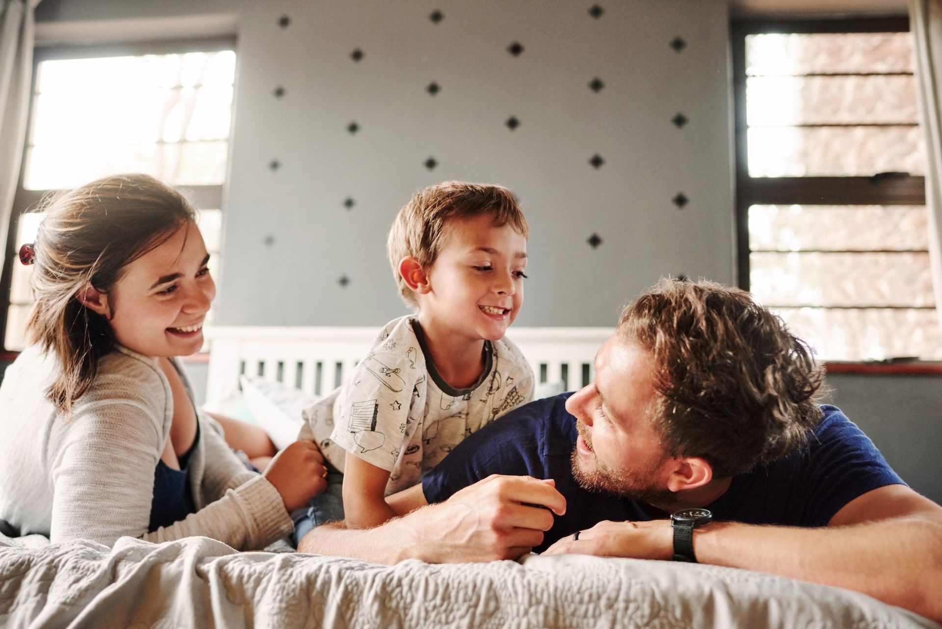 Three people lying on a bed together, smiling and interacting in a sunlit room with a grey polka-dot wall.