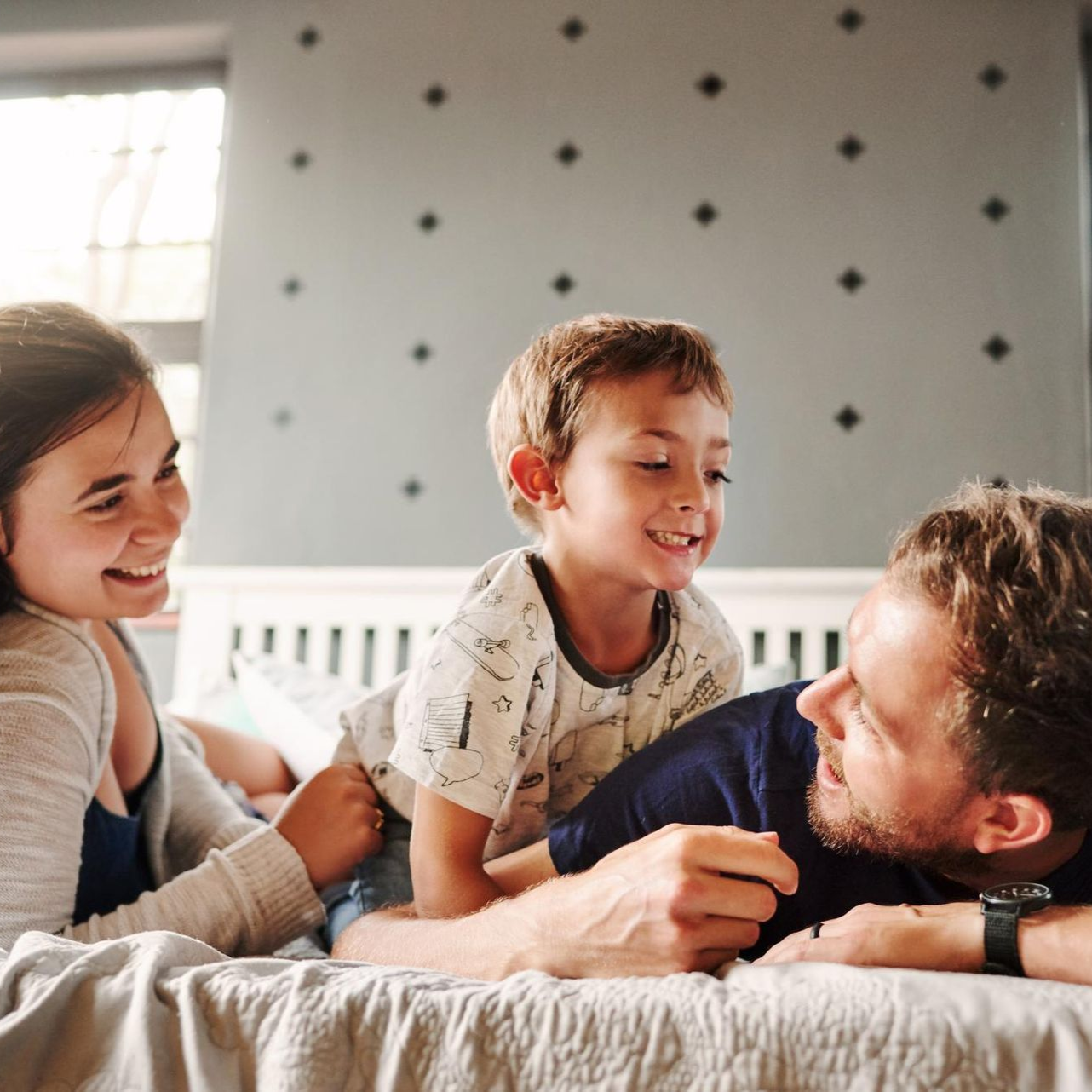 A family smiles and laughs together while relaxing on a bed in a room with a patterned grey wall.
