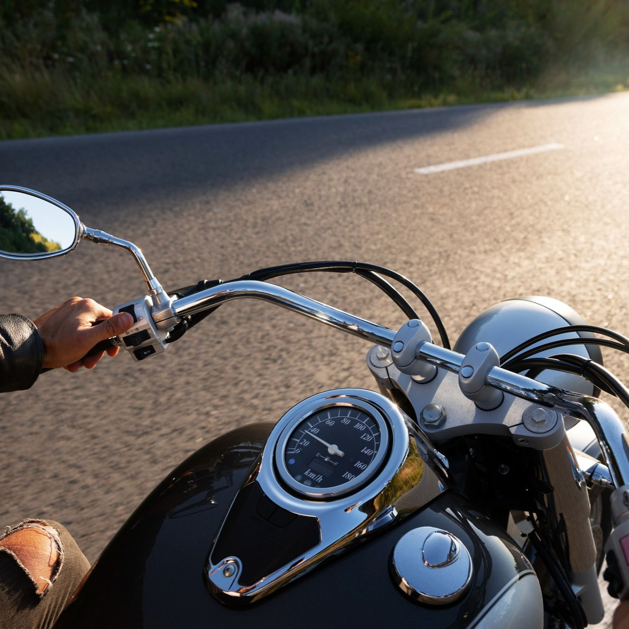 A person's hand grips the handlebar of a motorcycle while riding on a road in the sunlight.