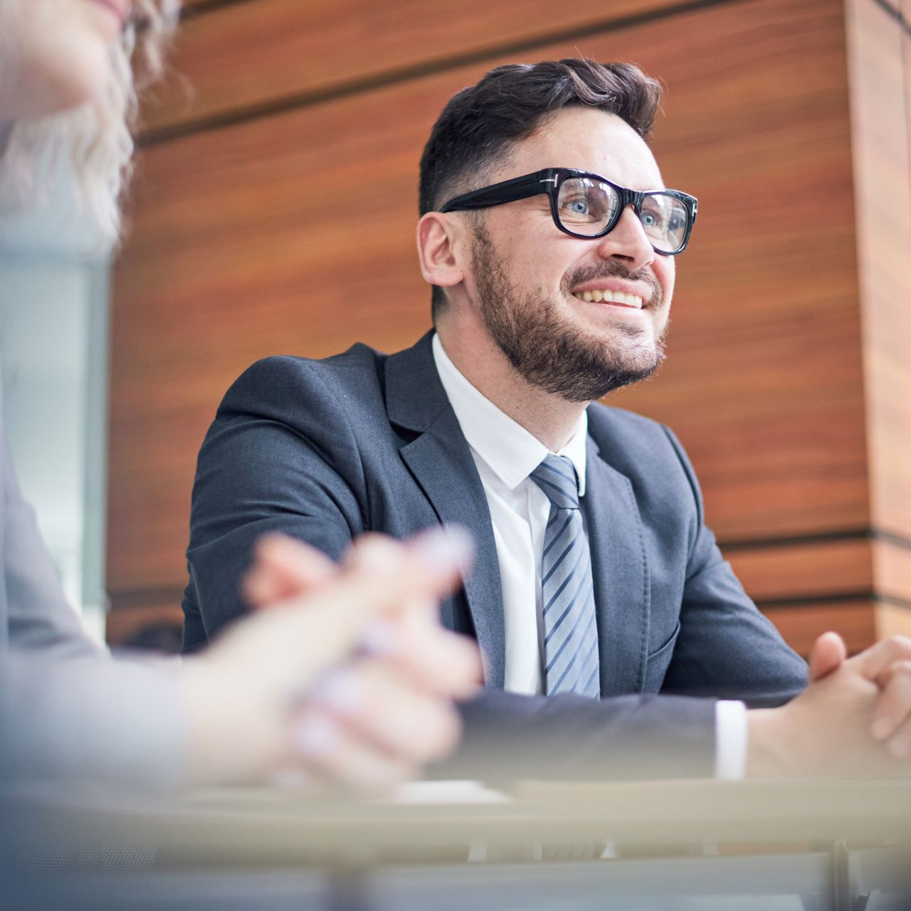 A man in a suit and glasses sits at a table, smiling while looking toward someone off-camera in a modern office setting.