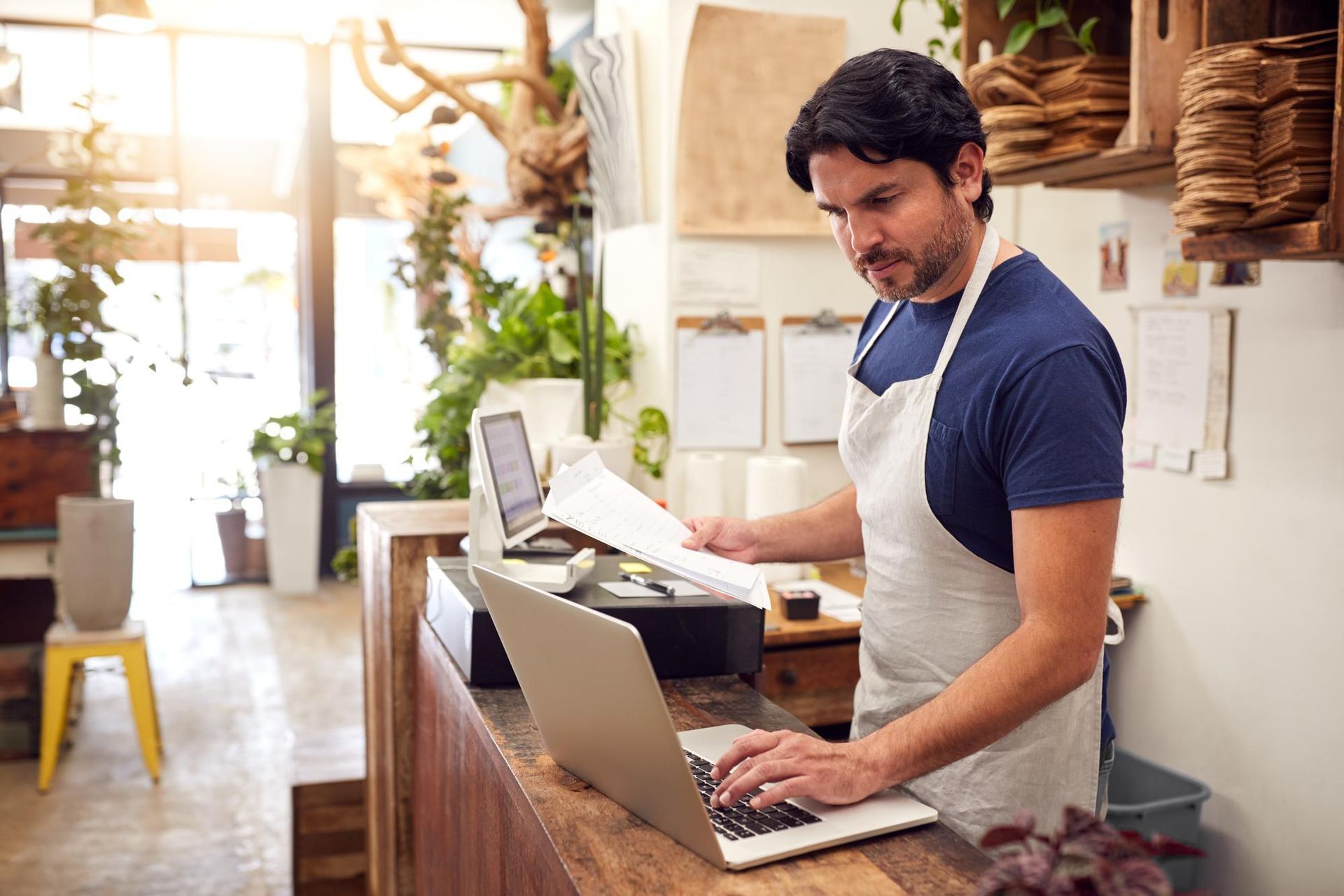 A person wearing an apron works on a laptop behind the wooden counter of a sunlit flower shop.