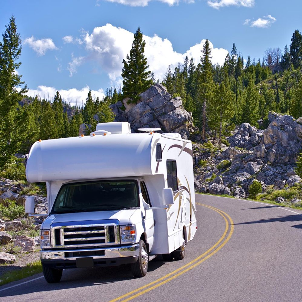 A white recreational vehicle driving along a paved road winding through a rocky, forested mountain landscape.