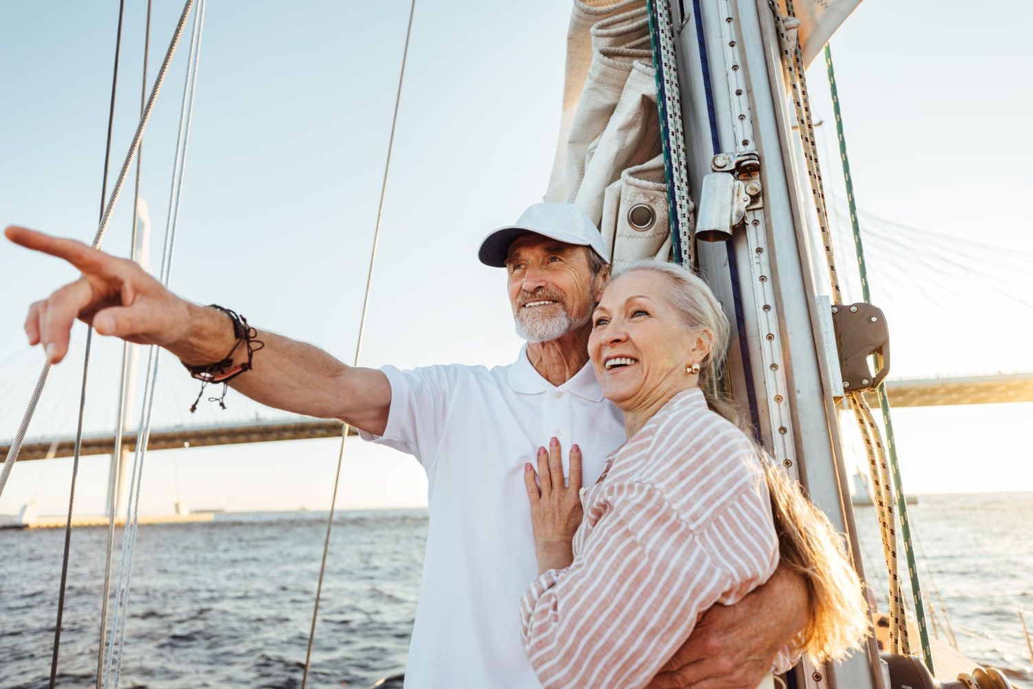 Two people on a sailboat, one pointing toward the horizon while the other smiles, with a bridge in the background.