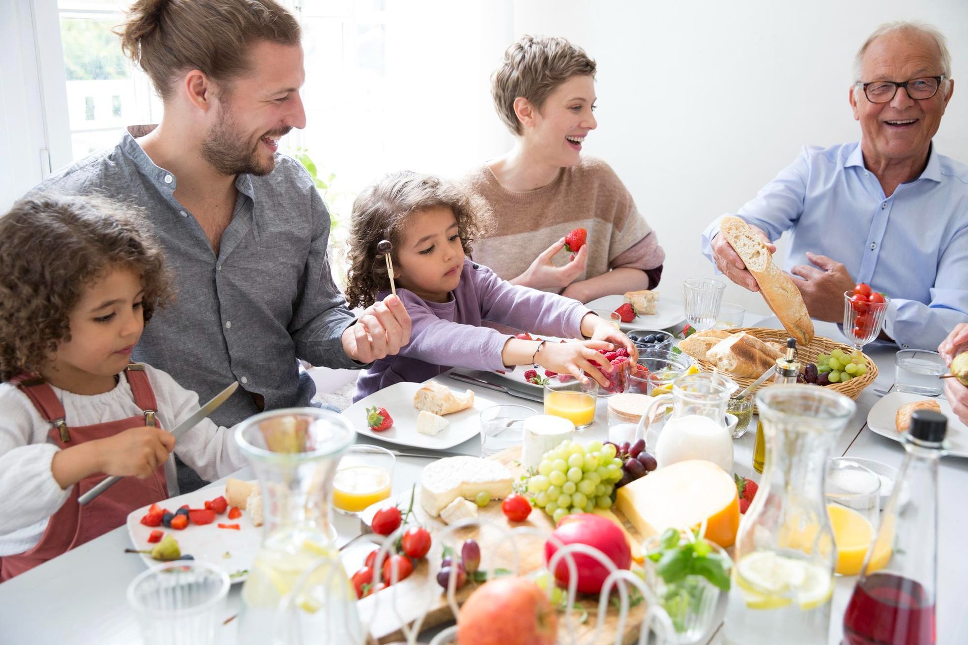 A family dining together at a bright table filled with bread, fruit, and drinks, all sharing a happy, casual moment.