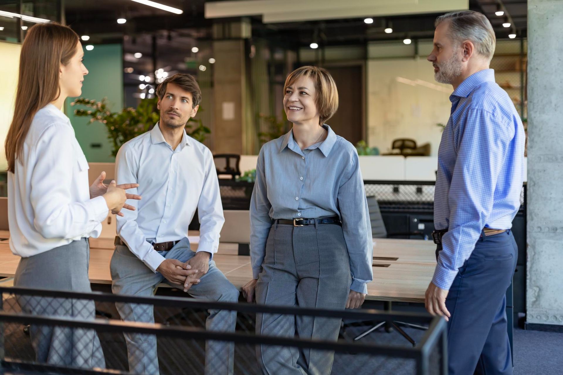 Four professionals stand in a modern, open-plan office space, talking and engaging in a collaborative business meeting.