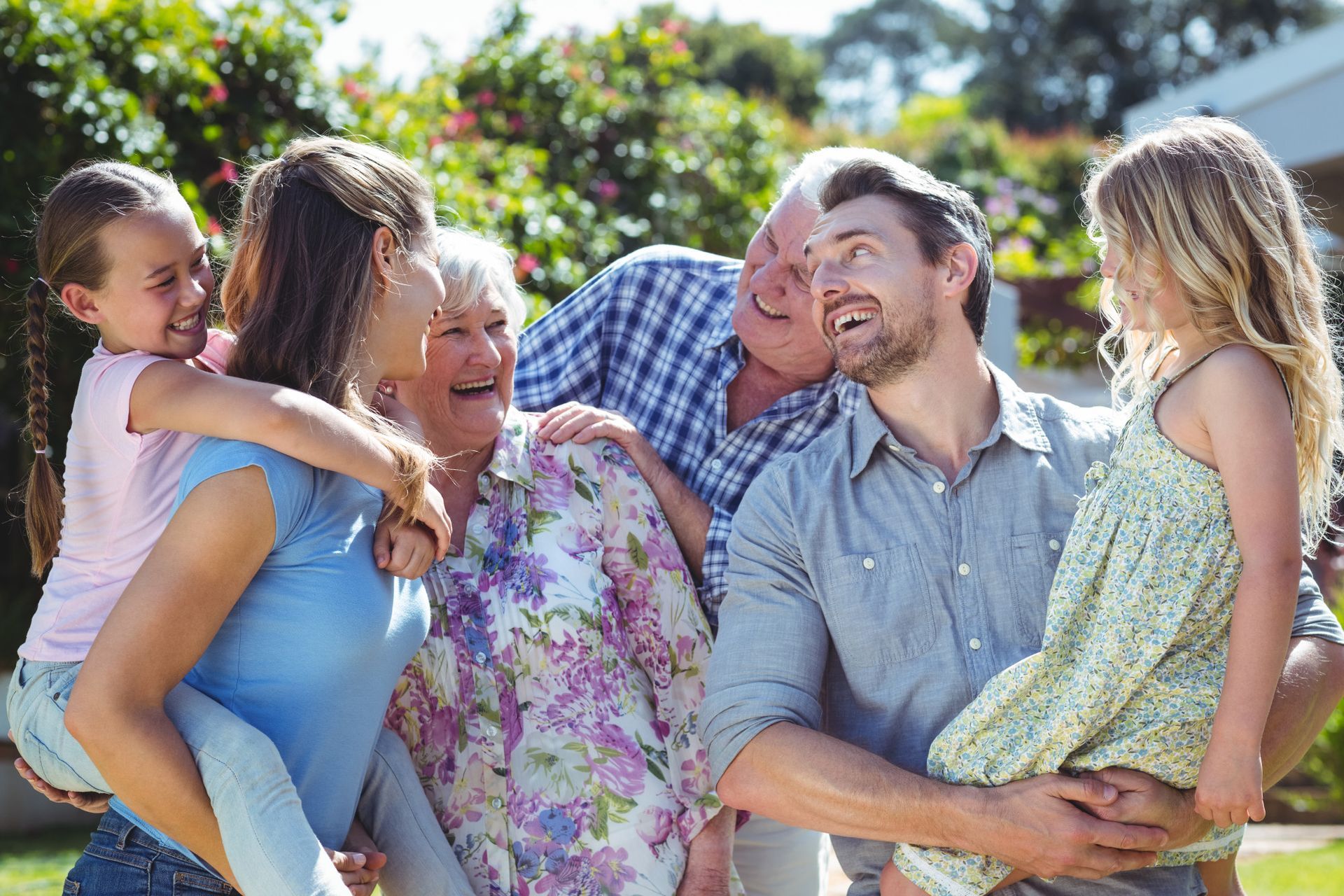 A multigenerational family smiles and interacts in an outdoor setting, with adults carrying two children on their backs.